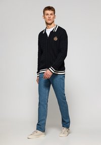 Cárdigan negro de punto trenzado con cremallera en el frente, cuello de rayas blancas y puños acanalados. Usado sobre una camiseta blanca y jeans azul claros.