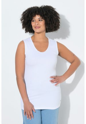 Kobieta o kręconych włosach, ubrana w białą, bezrękawnikową bluzkę i jasnoniebieskie dżinsy, stojąca z ręką na biodrze na białym tle.