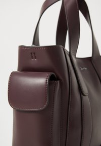 Bordo odinė rankinė su lygiu paviršiumi, turinti priekinį kišenę, elegantišką dizainą ir apvalius rankenėles. Siuvinėtos detalės suteikia elegancijos.