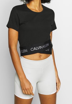 Kobieta ubrana w czarny krótki t-shirt Calvin Klein i białe, dopasowane szorty z wysokim stanem na białym tle.