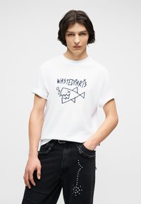 Νέος με μεσαίου μήκους σκούρα μαλλιά φοράει λευκό μπλουζάκι με το κείμενο "WASTEDPARIS" και ένα σχέδιο με στυλιζαρισμένο ψάρι, συνδυασμένο με μαύρο τζιν.