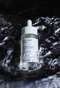 Klar glassflaske med hvit dråpetrekork, merket "mid/night hair serum." Væsken inni ser svakt grønnaktig ut, omgitt av vann.