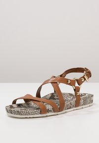 Brown leather sandal with multiple cross straps, adjustable ankle strap, and textured, scaly patterned sole with light gray bottom.