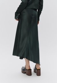 Μακριά πράσινη σατέν φούστα με ασύμμετρο τελείωμα, που χαρακτηρίζεται από ομαλή υφή και εφαρμοστή σιλουέτα, σε συνδυασμό με καφέ ξύλινα παπούτσια με πλατφόρμα.