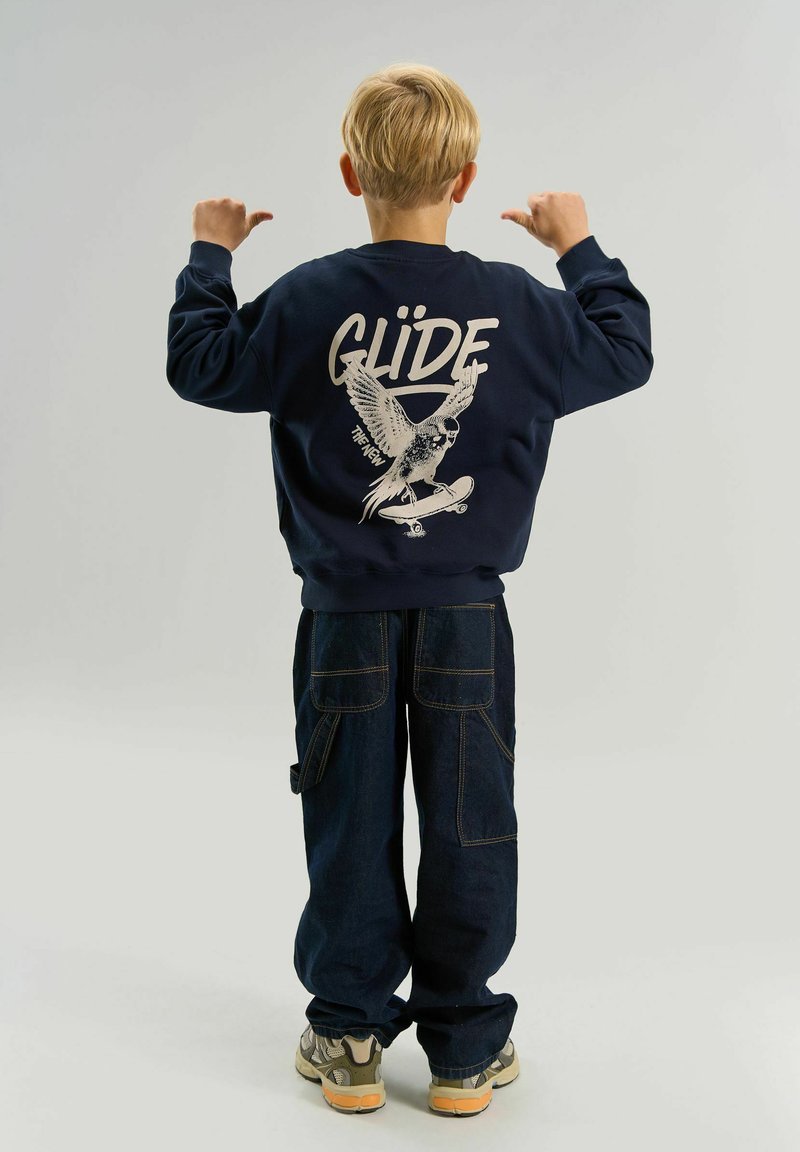 Enfant aux cheveux blonds montre du pouce le dos d'un sweat-shirt bleu marine avec un oiseau sur un skateboard et le mot « GLIDE ».