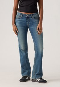 Jean flare bleu en denim lavé moyen avec des zones décolorées, design à cinq poches et coutures contrastées. Associés à des chaussures de sport noires.