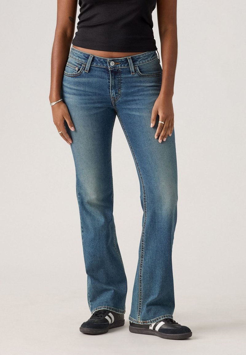 Jean flare bleu en denim lavé moyen avec des zones décolorées, design à cinq poches et coutures contrastées. Associés à des chaussures de sport noires.