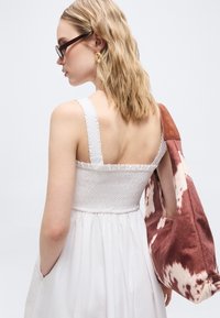 Γυναίκα με ξανθά κυματιστά μαλλιά που φορά γυαλιά ηλίου και χρυσά σκουλαρίκια, ντυμένη με λευκό αμάνικο φόρεμα, κρατώντας μια μεγάλη τσάντα με καφέ και λευκό σχέδιο.