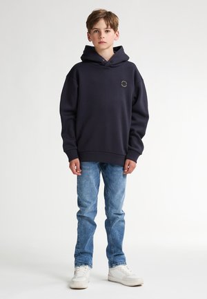 Sweat-shirt à capuche bleu marine avec un patch logo carré, porté avec un jean bleu clair et des baskets blanches. Design décontracté, texture douce.