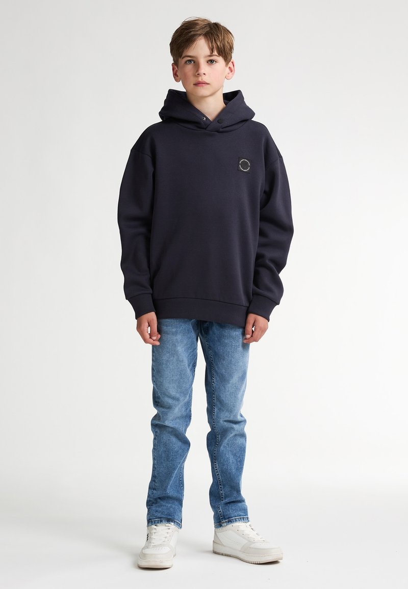 Felpa con cappuccio blu navy con un patch quadrato, abbinata a jeans azzurri e sneakers bianche. Design casual, texture morbida.