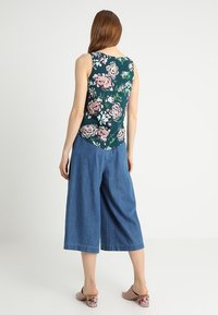 Débardeur floral en tissu vert foncé, associé à des culottes en denim larges. Présente des fleurs roses et blanches, un design décontracté et une coupe ample.