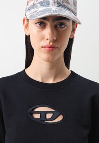 Sudadera negra con un detalle de logo recortado, combinada con una gorra estampada que presenta tonos de azul y rojo. Textura de tela suave evidente.