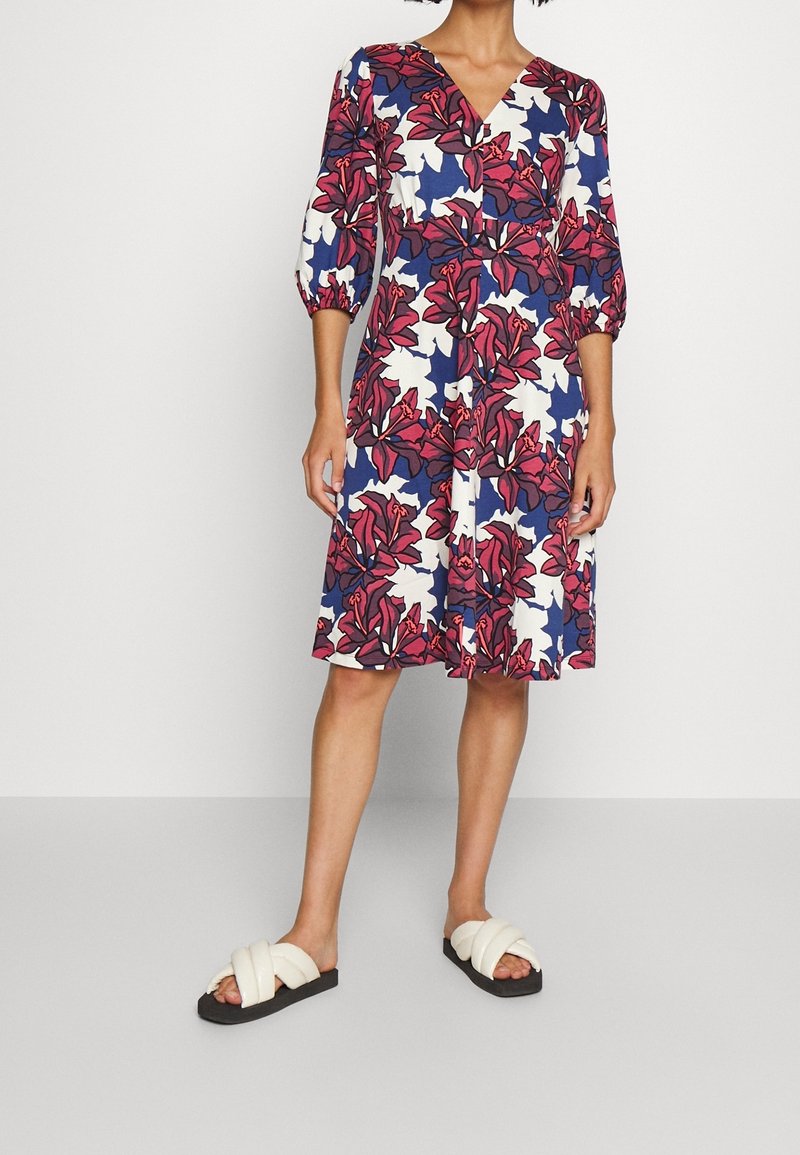 Vestido floral con escote en V que presenta flores rojas y blancas sobre un fondo azul, mangas abullonadas de tres cuartos y una silueta fluida en forma de A.