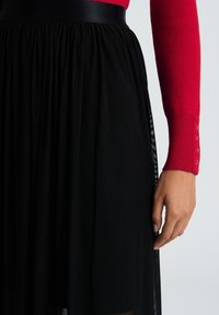 Svart plisserad kjol med smockad midja och satinmidja, i lager över ett genomskinligt tyg. Röd långärmad tröja synlig högst upp.