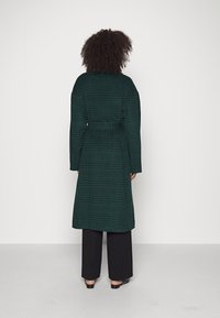 Cappotto lungo verde a quadri con cintura, caratterizzato da un taglio dritto e maniche ampie. Indossato sopra pantaloni neri e scarpe basse. Sfondo semplice.