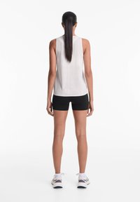 Bílý bezrukávový sportovní top s perforovanou texturou, v kombinaci s černými šortkami. Modelka má na sobě tenisky a vlasy stylizované do culíku.