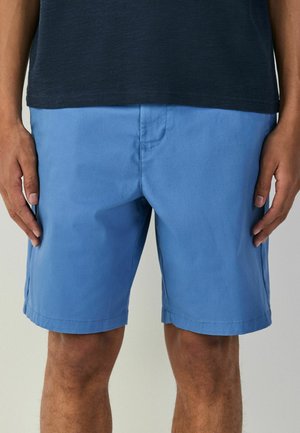 Bărbat purtând pantaloni scurți albastru deschis până la jumătatea coapsei și o cămașă bleumarin închis, stând cu mâinile relaxate pe lângă corp pe un fundal simplu.