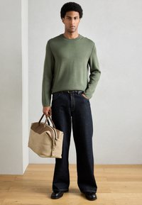 Camisola verde, jeans azul escuro de perna larga e uma bolsa bege com detalhes castanhos. Chão de madeira e parede cinza clara como fundo.