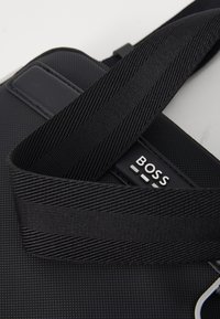 Black crossbody bag made of textured material. Features a logo, adjustable strap, and smooth accents. Minimalist design with functional details.