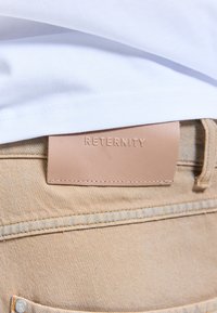 Patch en cuir fauve embossé du mot « RETERNITY » sur la ceinture d'un jean beige sous l'ourlet d'une chemise blanche.