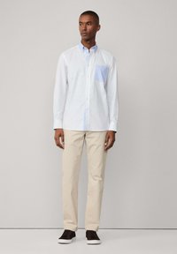 Camicia bianca con tasca azzurra chiara, abbinata a pantaloni beige e sneaker marrone scuro, su uno sfondo neutro.