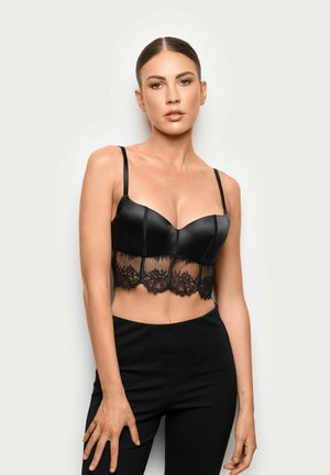 Joven mujer con cabello liso lleva un bralette de satén negro con borde de encaje y pantalones negros de talle alto sobre un fondo claro y sencillo.
