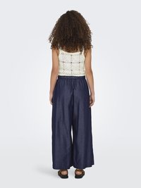 Witte gehaakte tanktop met vierkant patroon, gecombineerd met losse navy wijde broek. Model staat met de rug naar voren, toont de details van de outfit.