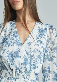 Blumenkleid mit weißem Grund und blauen Blüten sowie Kolibris. V-Ausschnitt-Design mit Puffärmeln und einem Stoffgürtel.