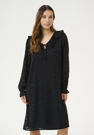 Schwarzes Kleid mit langen Ärmeln, Rüschenausschnitt, Schlüssellochdetail und strukturiertem Wellenmuster. Hergestellt aus leichtem, transparentem Stoff.
