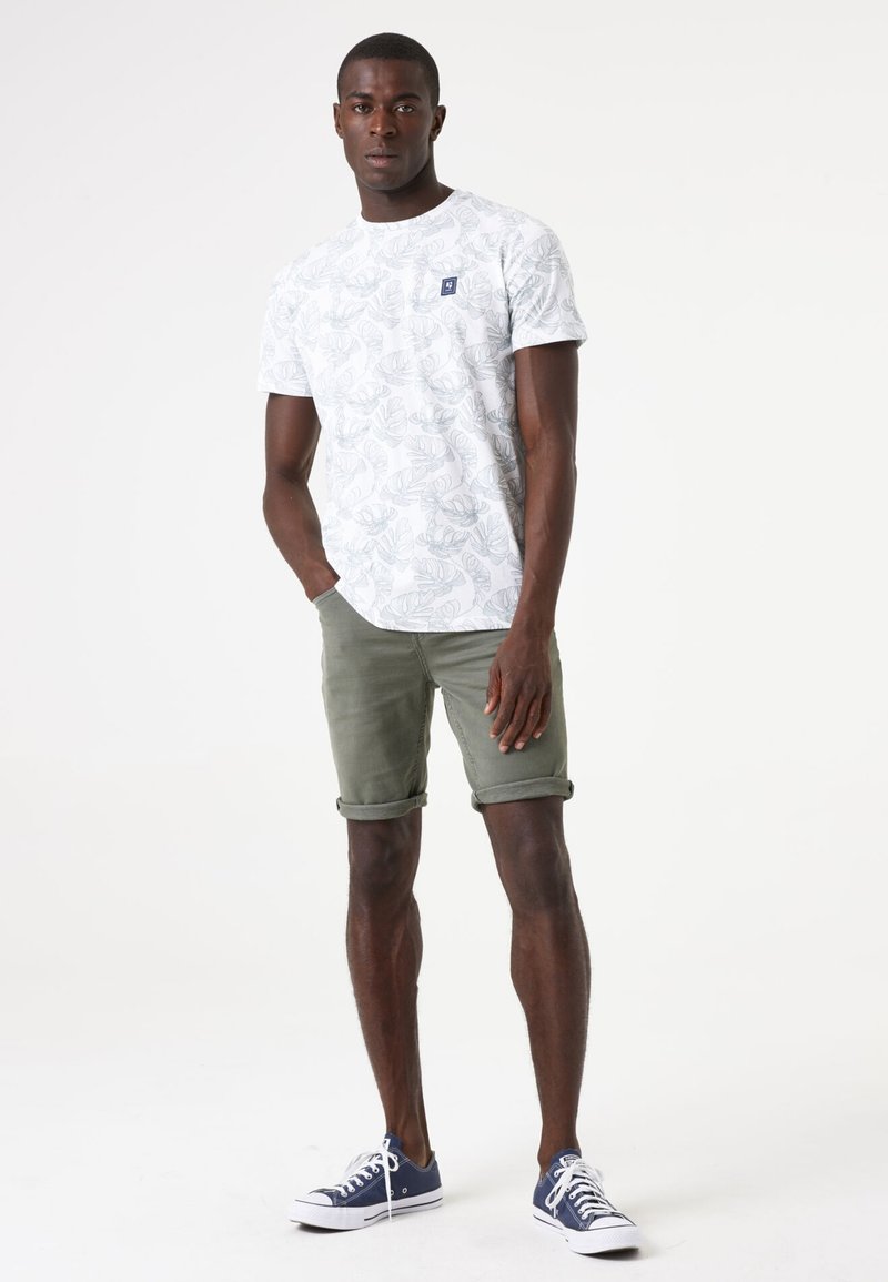 Witte t-shirt met korte mouwen en een groen bladerenpatroon, gecombineerd met olijfgroene opgerolde shorts en marineblauwe sneakers. Heeft een klein logo-detail.
