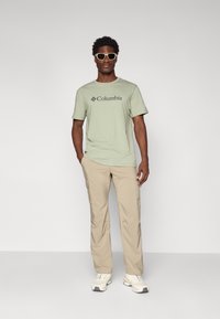 Light green short-sleeve t-shirt with "Columbia" logo, beige cargo pants, and white athletic shoes. Model wearing white sunglasses.