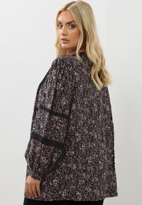 Blusa nera con un motivo floreale in rosa e bianco. Presenta maniche lunghe a sbuffo e dettagli testurizzati neri sulle spalle. Vestibilità morbida.