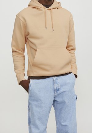 Man met beige capuchontrui met voorzak en lichtblauwe spijkerbroek, handen in zakken, tegen een effen lichte achtergrond.