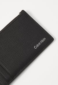 Zwarte textuurportemonnee van leer; heeft meerdere kaartsleuven en een zilveren "Calvin Klein" logo aan de voorkant.