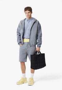 Junger Mann trägt blaues Hemd, Jeansjacke und Shorts, gelbe Turnschuhe, trägt schwarze gesteppte Tragetasche, steht vor weißem Hintergrund.