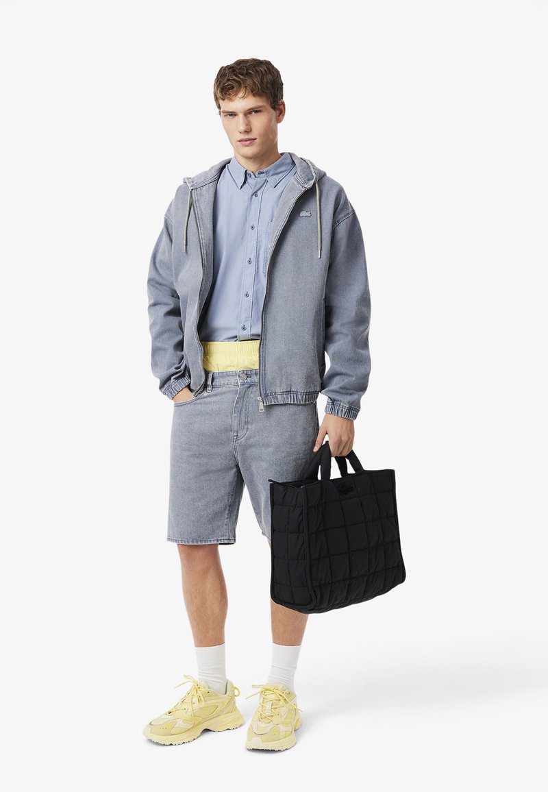 Junger Mann trägt blaues Hemd, Jeansjacke und Shorts, gelbe Turnschuhe, trägt schwarze gesteppte Tragetasche, steht vor weißem Hintergrund.