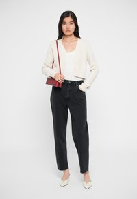 Avkortet lys beige cardigan med knapper, hvit tanktop, høyt liv svarte jeans og kremfargede spisse sko; rød håndveske over skulderen.