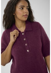 Pull en tricot à manches courtes de couleur violet profond avec un col, comportant cinq boutons ronds de couleur claire sur le devant et une texture douce.
