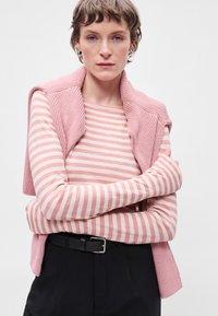 Colete de malha rosa sobre uma blusa de manga longa com riscas rosa e brancas. A modelo veste calças pretas de cintura alta e um cinto preto, com os braços cruzados.