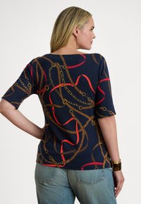 Žena se světlými vlasy, která má na sobě tmavě modrou košili s potiskem hnědých a červených řetězů a lan, doplněnou světle modrými džínami, otočená zády.