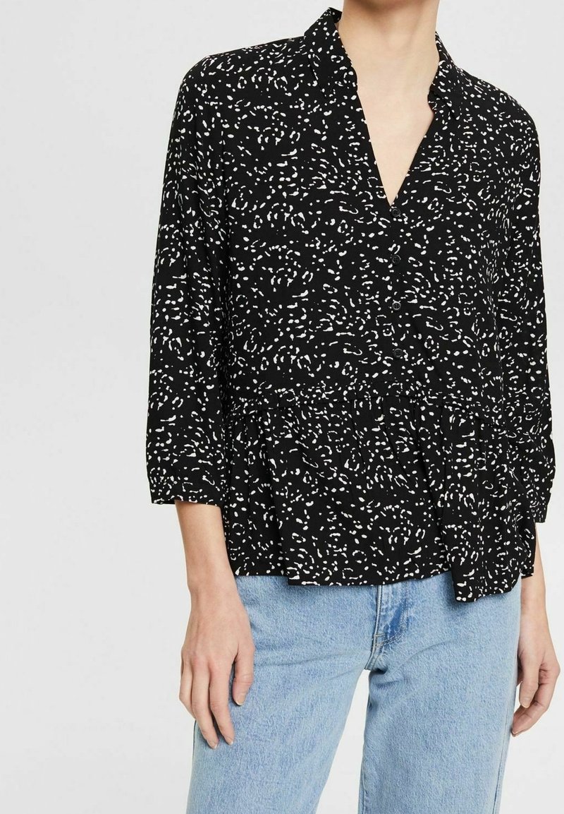 Personne portant un chemisier noir boutonné avec des taches abstraites blanches et un jean bleu clair, vu du cou jusqu'aux hanches.