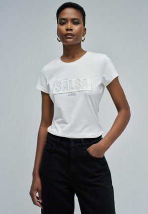 Mujer con pelo corto que lleva pendientes de aro dorados, camiseta blanca de "SALSA JEANS" y pantalones negros con una mano en el bolsillo, contra un fondo liso.