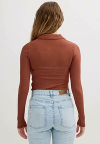 Camisa de manga larga de color óxido con cuello, combinada con jeans de tiro alto de color azul claro que presentan bolsillos traseros prominentes y una etiqueta.