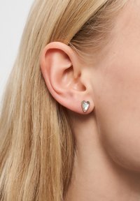 Blont hår bakom vänster öra, bär en liten hjärtformad silverspik med en klar ädelsten.