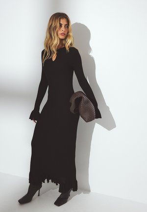 Lange zwarte geribbelde jurk met een keyhole halslijn, gerimpelde zoom en uitlopende mouwen. Gecombineerd met een gestructureerde bruine clutch en hoge laarzen.