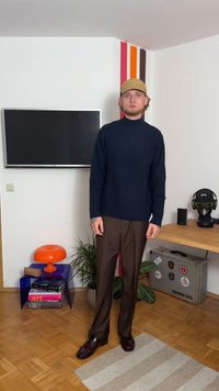 Mornarsko pleteno pulover s visokim ovratnikom, rjave hlače in temni čevlji. Dodatki vključujejo svetlo kapo. Preprost, teksturiran dizajn in sproščen kroj.