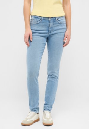 Világoskék slim fit farmer, fehér tornacipővel és világos sárga felsővel viselve, egy sima háttér előtt álló személyen.