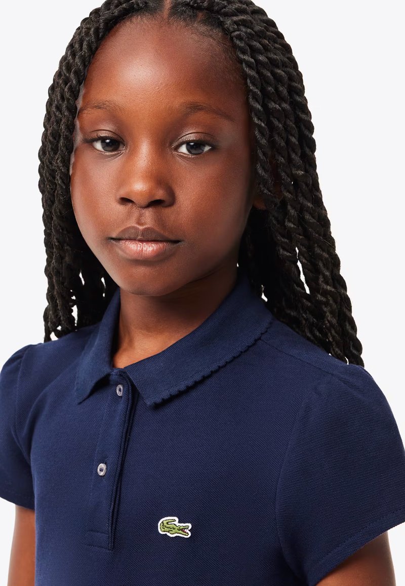 Mörkblå polo t-shirt med korta puffärmar, ribbad krage och en liten grön broderad logotyp på vänster bröst. Bomullstextur.