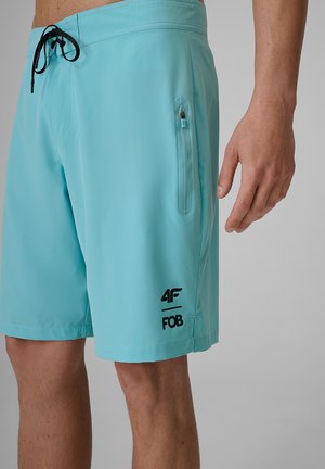 Mężczyzna noszący jasnoniebieskie spodenki kąpielowe z czarnym sznurkiem i zapinaną na zamek kieszenią boczną, z logo "4F FOB" na lewej nodze.