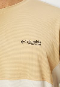Columbia Titanium srajca, strukturirana svetlo bež tkanina, okrogla izrezana vrata, z črnim logotipom na prsih, gladek bel spodnji del.